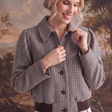 A woman models a brown and beige checkered cropped jacket with dark brown buttons and a matching dark brown belt, paired with a light beige wrap skirt, set against a scenic landscape backdrop.
