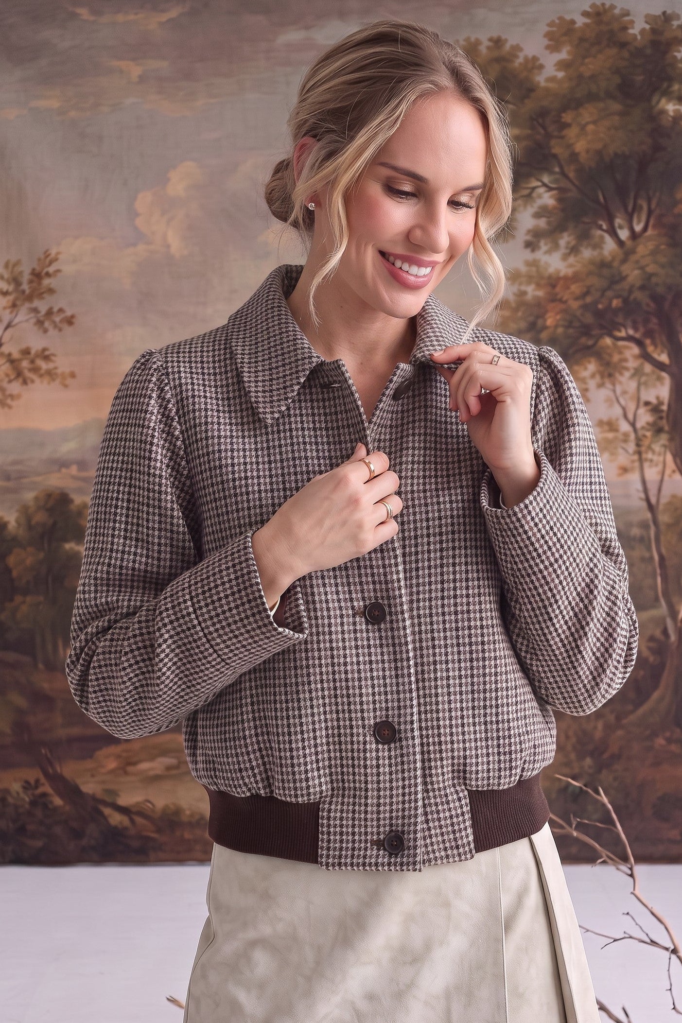 A woman models a brown and beige checkered cropped jacket with dark brown buttons and a matching dark brown belt, paired with a light beige wrap skirt, set against a scenic landscape backdrop.