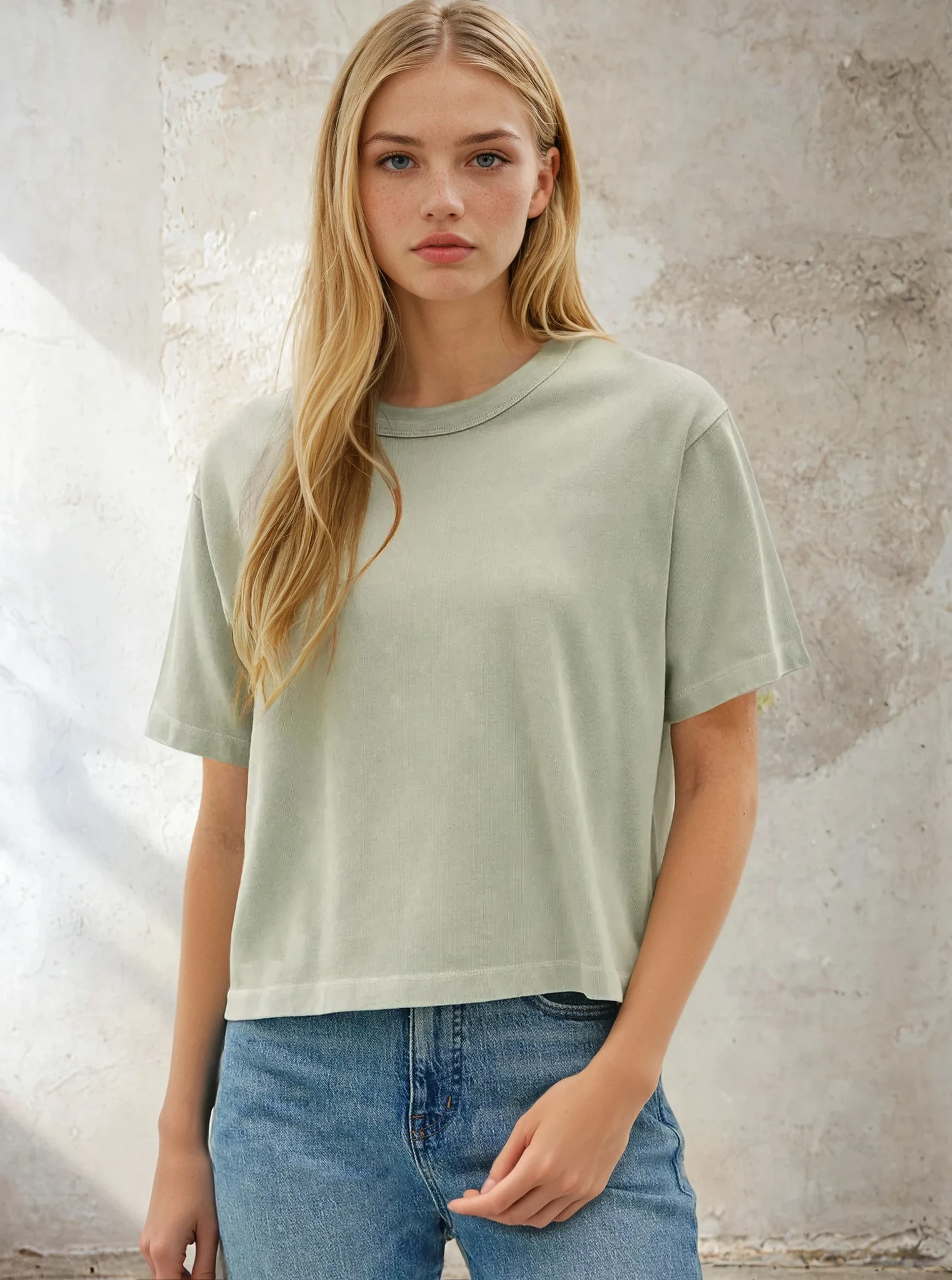Woman wearing a light green t-shirt and blue jeans against a textured wall.