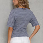 Woman wearing a blue striped shirt and white pants against a gray wall