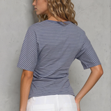 Woman wearing a blue striped shirt and white pants against a gray wall