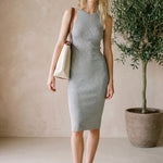 Woman in a gray dress standing next to a potted plant in a minimalistic room.