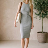 Woman in a gray dress standing next to a potted plant in a minimalistic room.