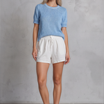 Woman wearing a blue sweater and white shorts against a gray concrete wall.