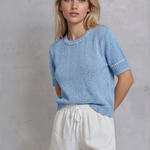 Woman wearing a light blue sweater and white shorts against a gray concrete wall.