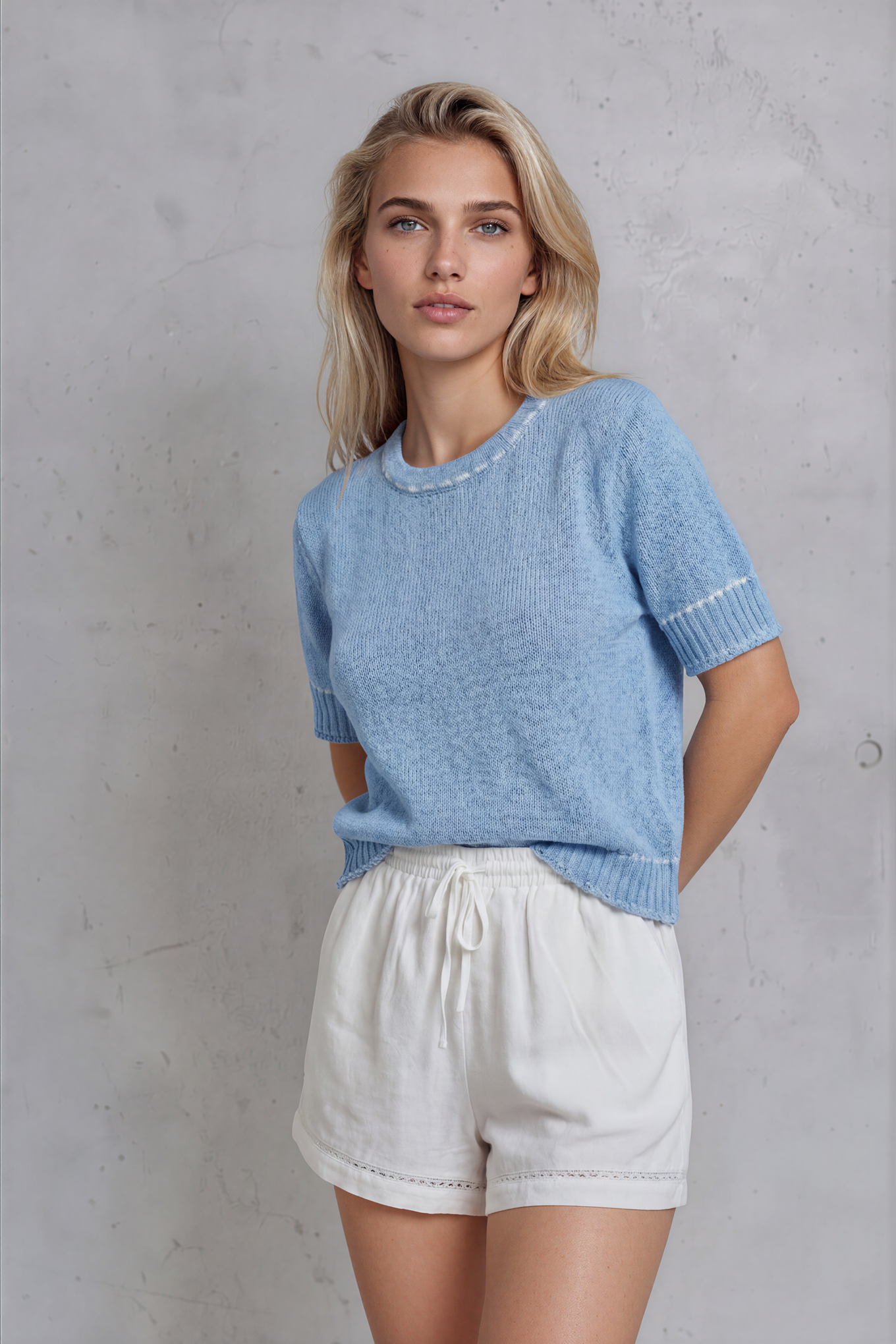 Woman wearing a light blue sweater and white shorts against a gray concrete wall.
