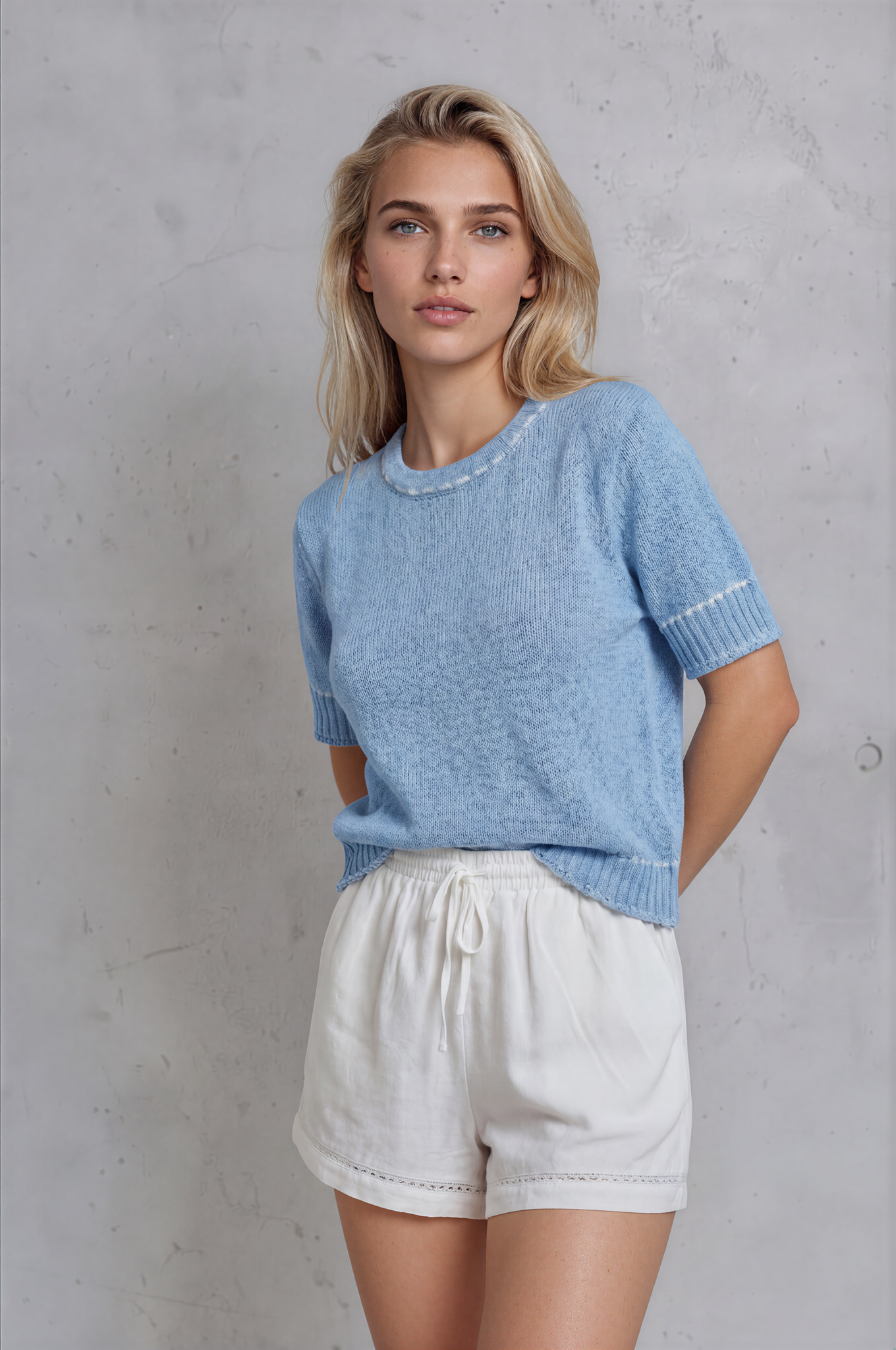 Woman wearing a light blue sweater and white shorts against a gray concrete wall.