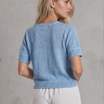Woman wearing a blue knitted top and white shorts against a gray concrete wall.
