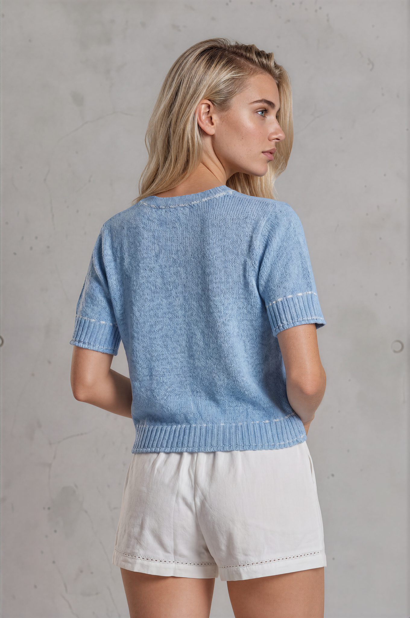 Woman wearing a blue knitted top and white shorts against a gray concrete wall.