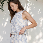 Woman wearing a white floral dress standing against a white wall with greenery.