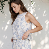 Woman wearing a white floral dress standing against a white wall with greenery.