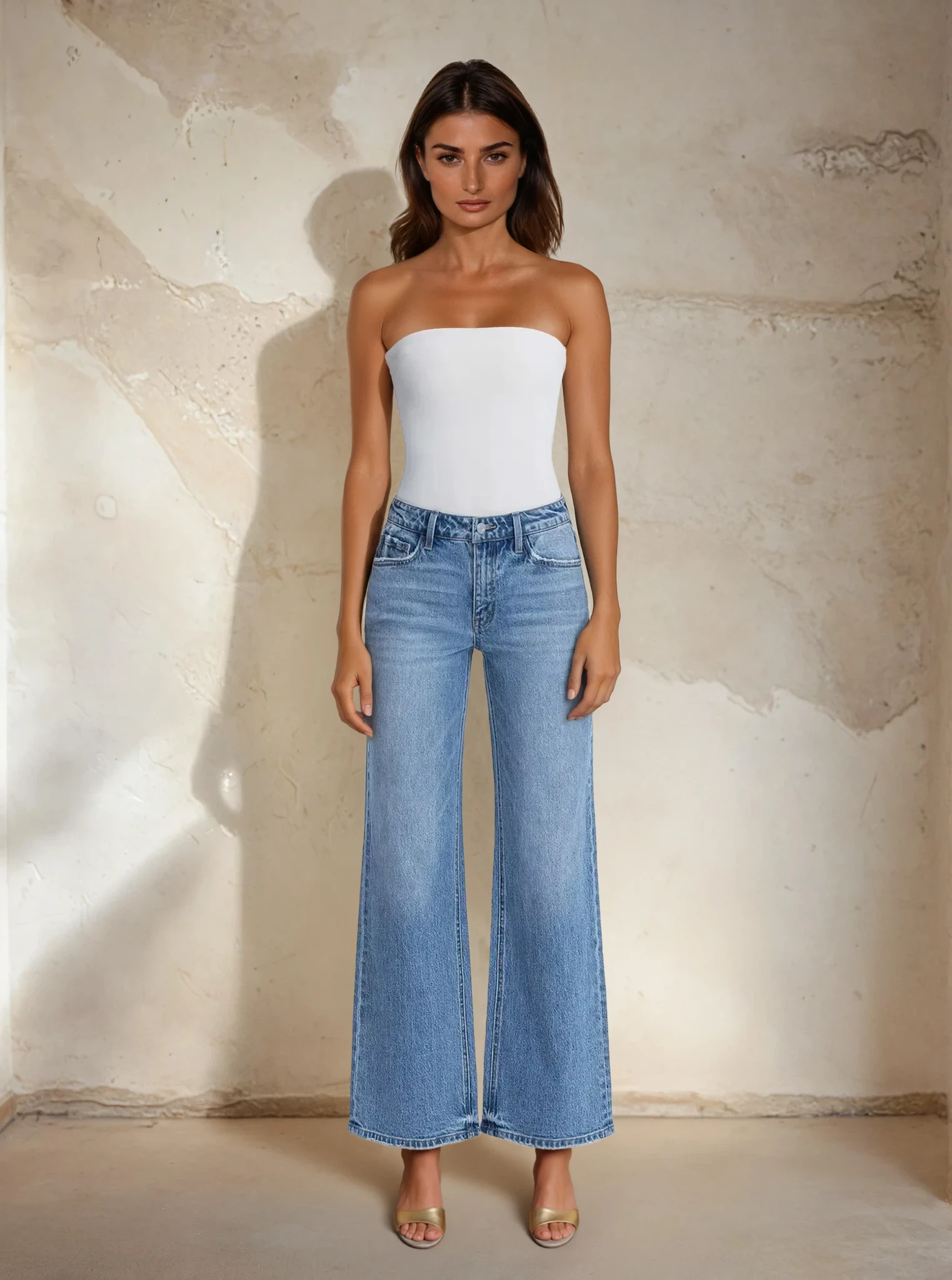 Woman wearing a white strapless top and blue jeans against a textured beige wall.