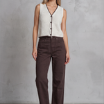 Woman wearing a white sleeveless top with black buttons and brown pants against a gray concrete wall.