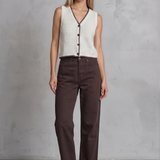 Woman wearing a white sleeveless top with black buttons and brown pants against a gray concrete wall.