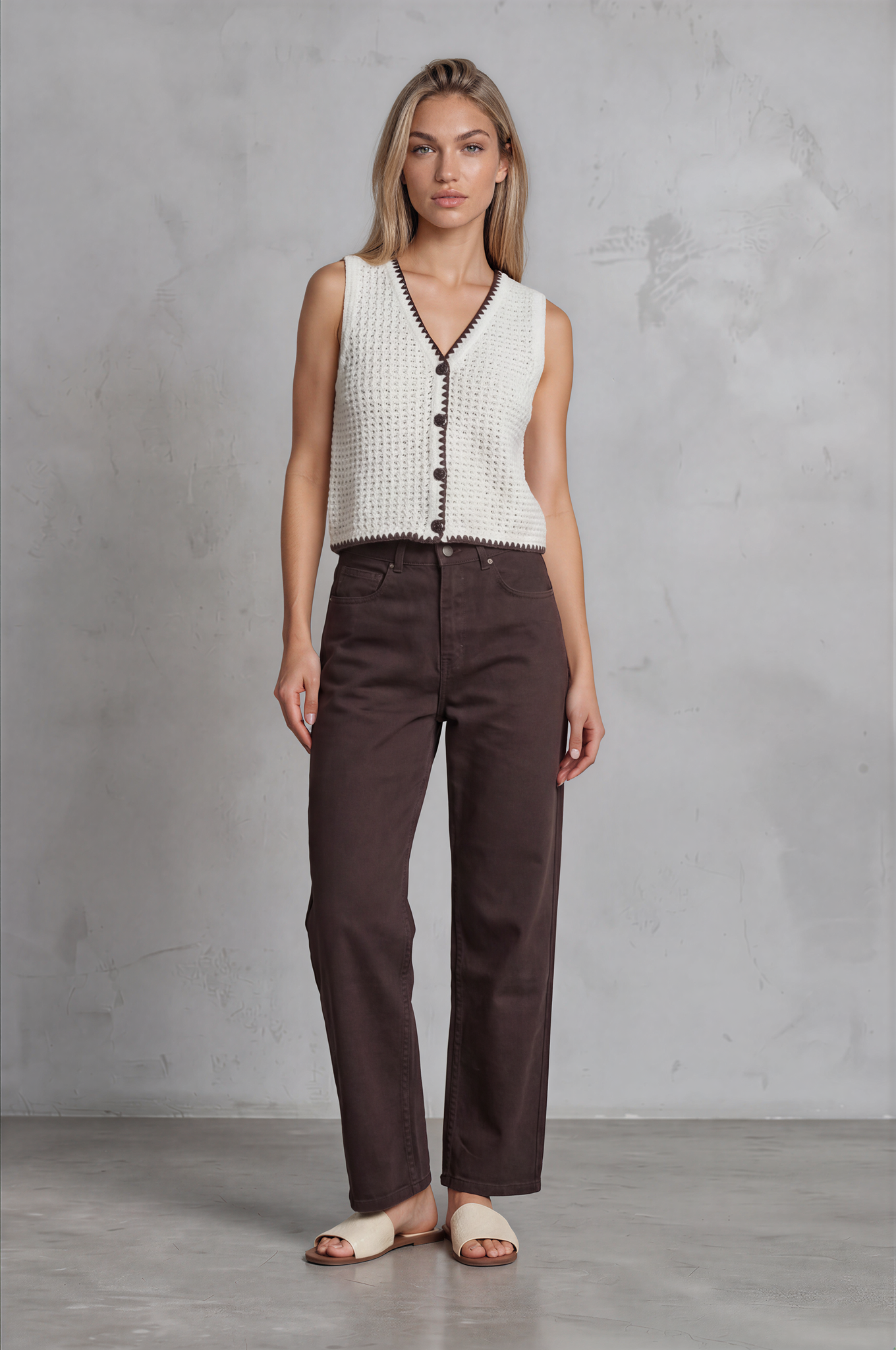 Woman wearing a white sleeveless top with black buttons and brown pants against a gray concrete wall.