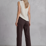 Woman wearing a white sleeveless top and brown pants against a gray concrete wall.