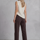 Woman wearing a white sleeveless top and brown pants against a gray concrete wall.