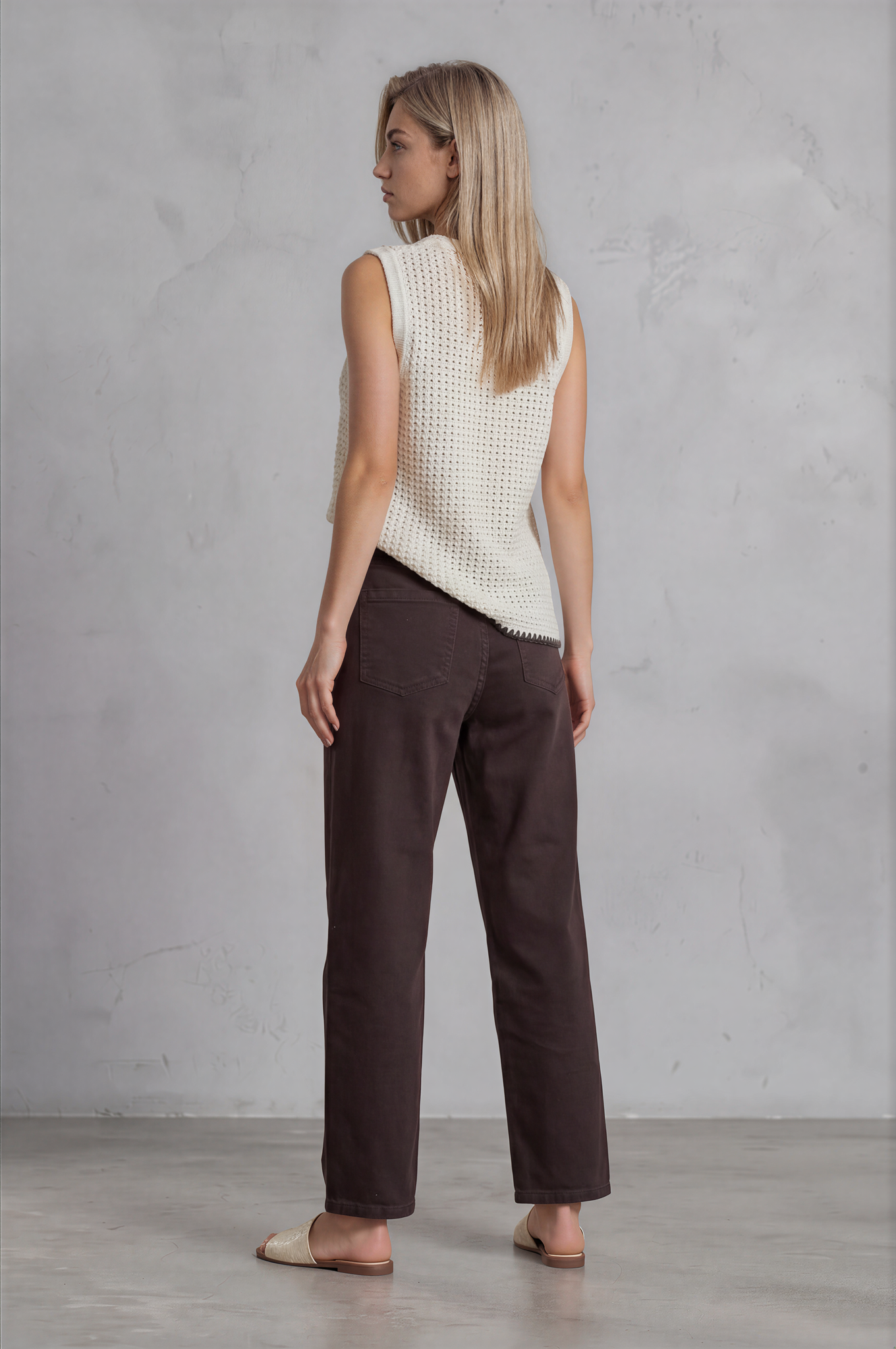 Woman wearing a white sleeveless top and brown pants against a gray concrete wall.