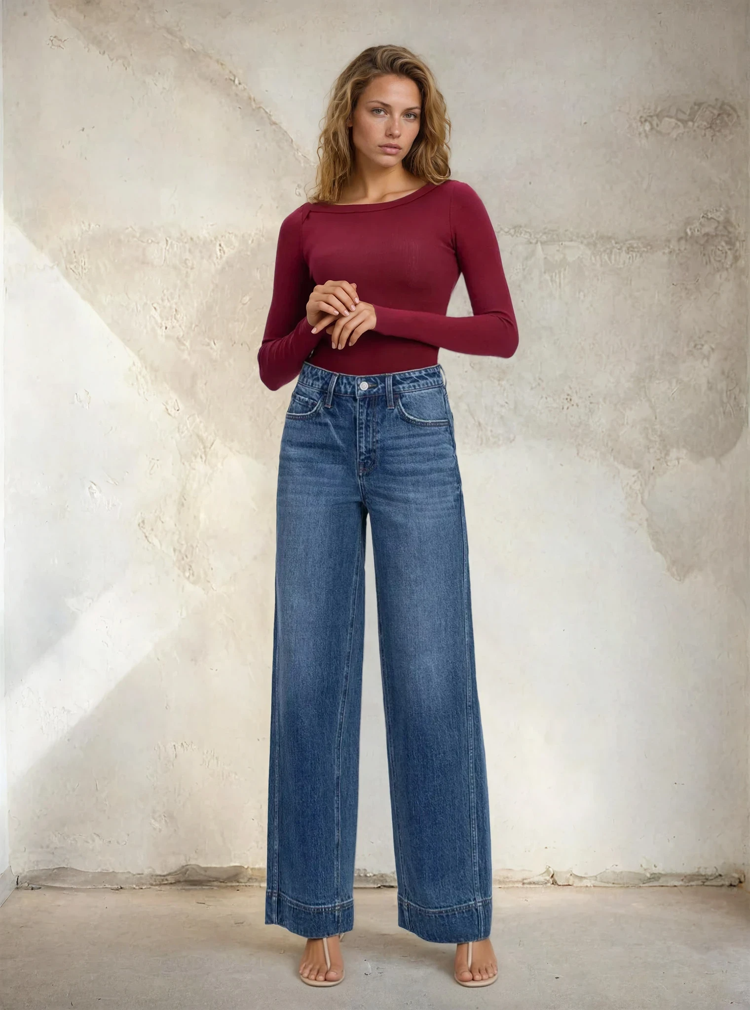 Woman wearing a burgundy top and blue jeans against a textured wall.