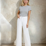 Woman wearing a gray ribbed t-shirt and white wide-leg pants against a textured wall.