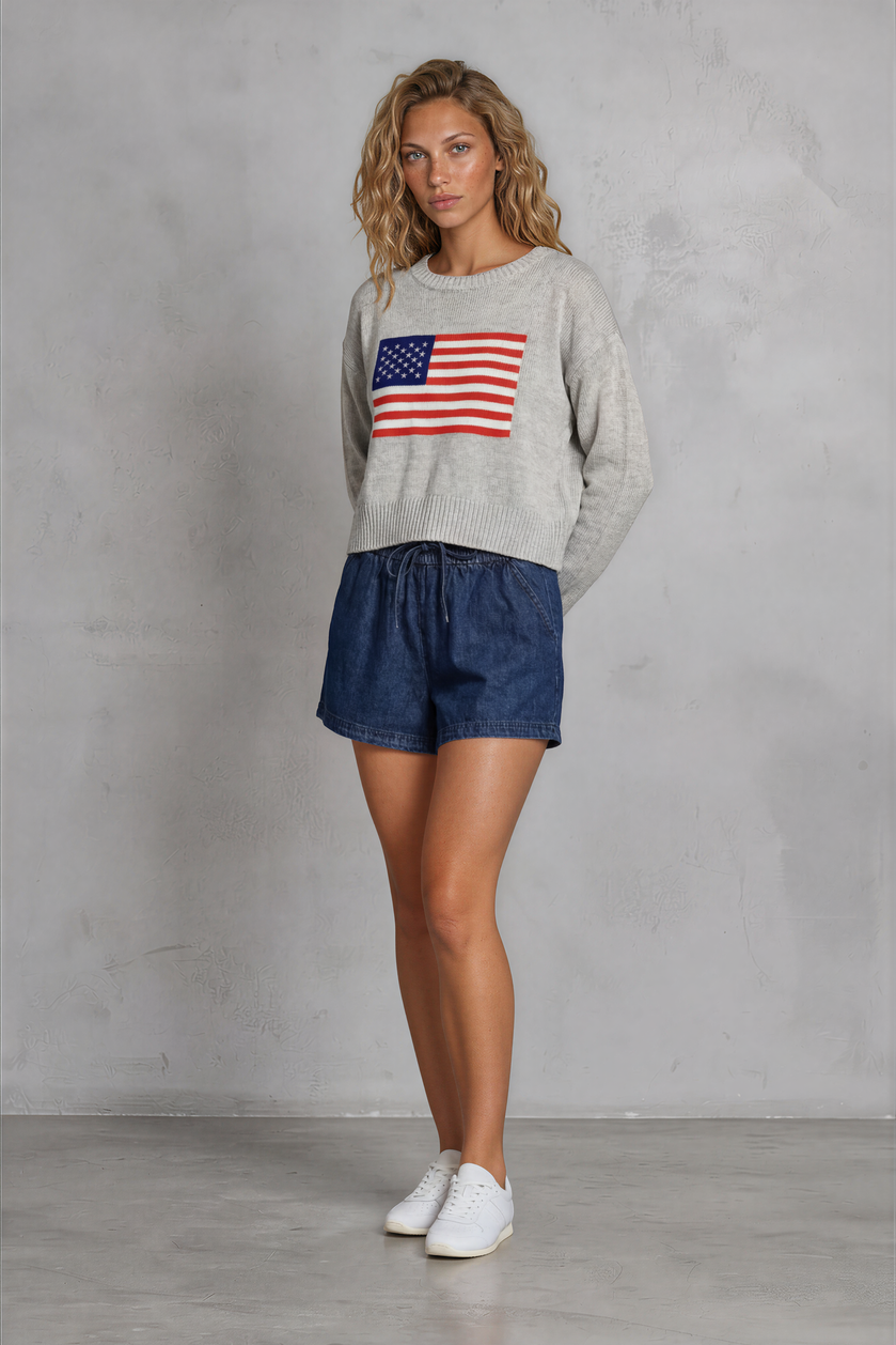 Woman wearing a sweater with an American flag design and denim shorts against a gray background