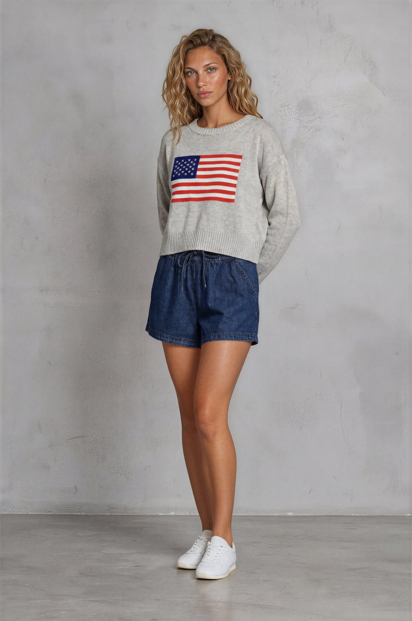Woman wearing a sweater with an American flag design and denim shorts against a gray background