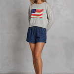 Woman wearing a sweater with an American flag design and denim shorts against a gray background