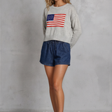 Woman wearing a sweater with an American flag design and denim shorts against a gray background