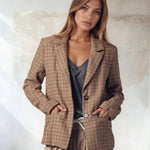 Woman wearing a plaid blazer over a gray top and beige pants against a textured wall.