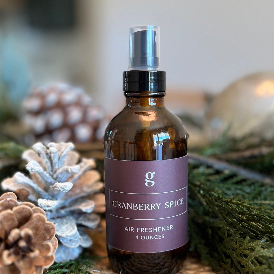 A spray bottle labeled 'Graced Cranberry Spice Air Freshener' placed on a table with pine cones and greenery in the background.
