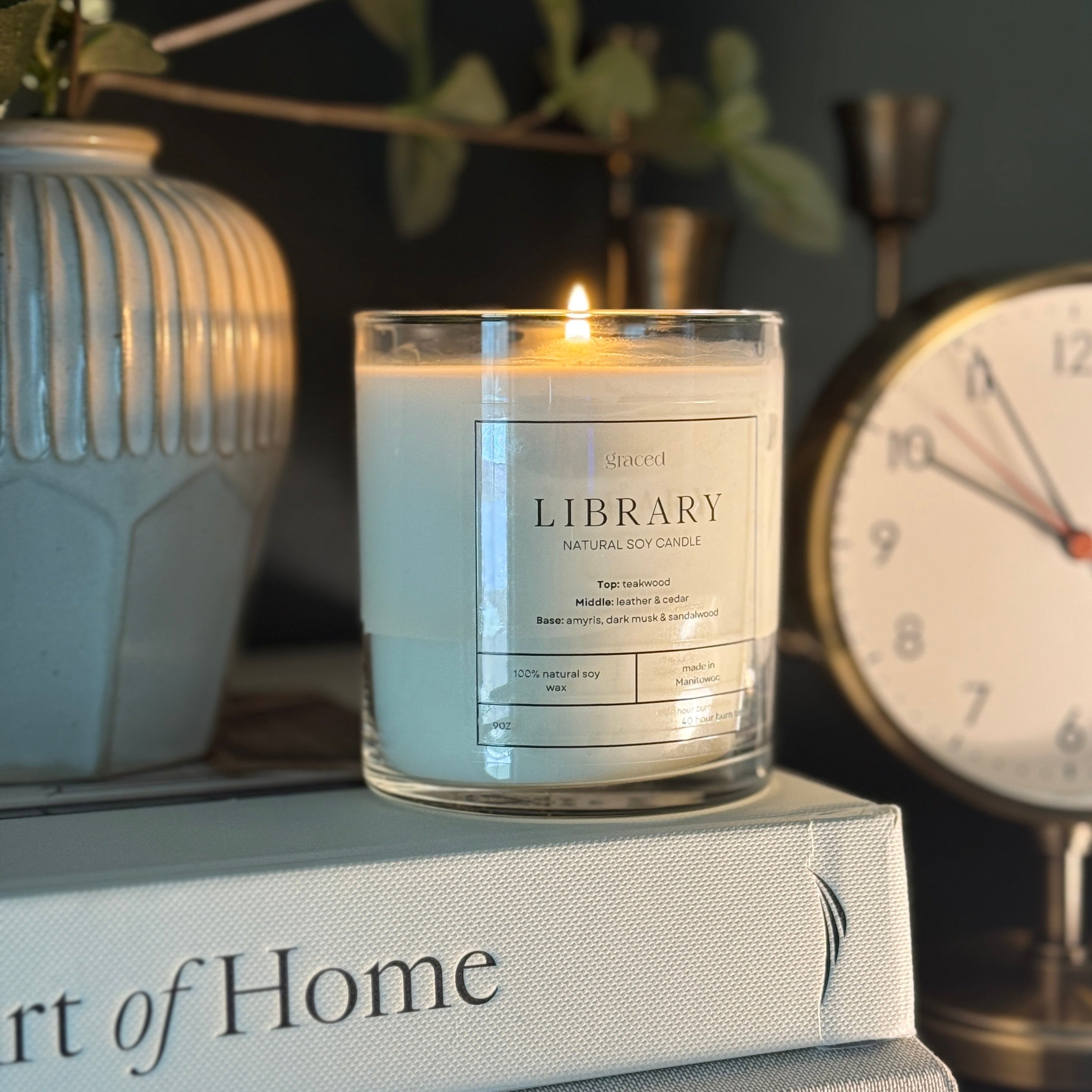 Candle labeled 'Library' on books with a clock and plant in the background