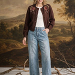 A woman stands in front of a classical landscape painting backdrop, wearing a dark brown embroidered cardigan over a white t-shirt, light-wash wide-leg jeans, and brown suede loafers.