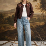 A woman stands in front of a classical landscape painting backdrop, wearing a dark brown embroidered cardigan over a white t-shirt, light-wash wide-leg jeans, and brown suede loafers.