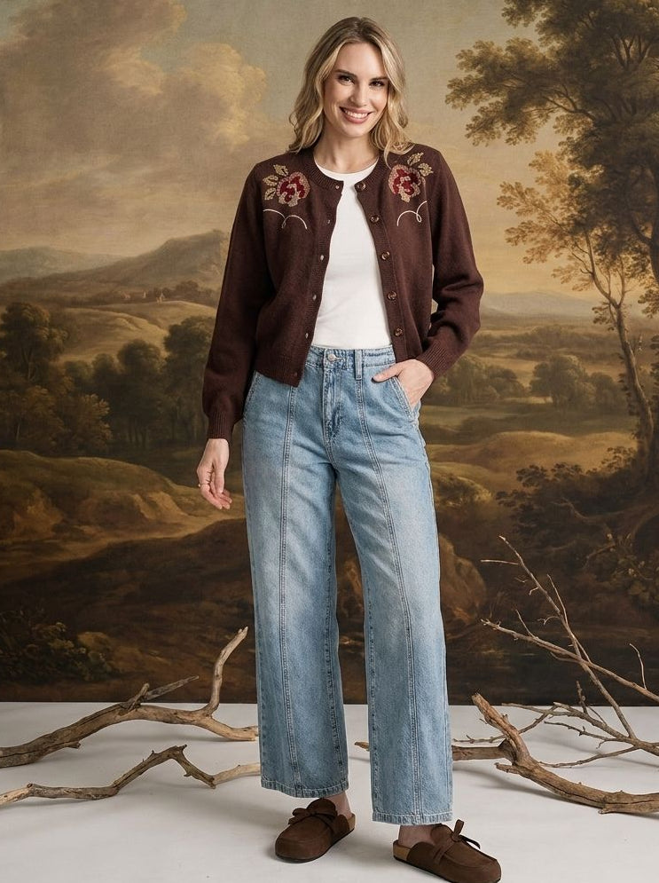 A woman stands in front of a classical landscape painting backdrop, wearing a dark brown embroidered cardigan over a white t-shirt, light-wash wide-leg jeans, and brown suede loafers.