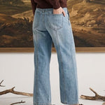A woman stands in front of a large framed landscape painting, wearing a dark brown long-sleeve top, light blue wide-leg jeans, and brown slip-on shoes.