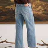 A woman stands in front of a large framed landscape painting, wearing a dark brown long-sleeve top, light blue wide-leg jeans, and brown slip-on shoes.