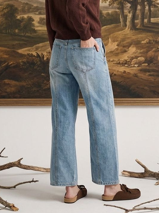 A woman stands in front of a large framed landscape painting, wearing a dark brown long-sleeve top, light blue wide-leg jeans, and brown slip-on shoes.