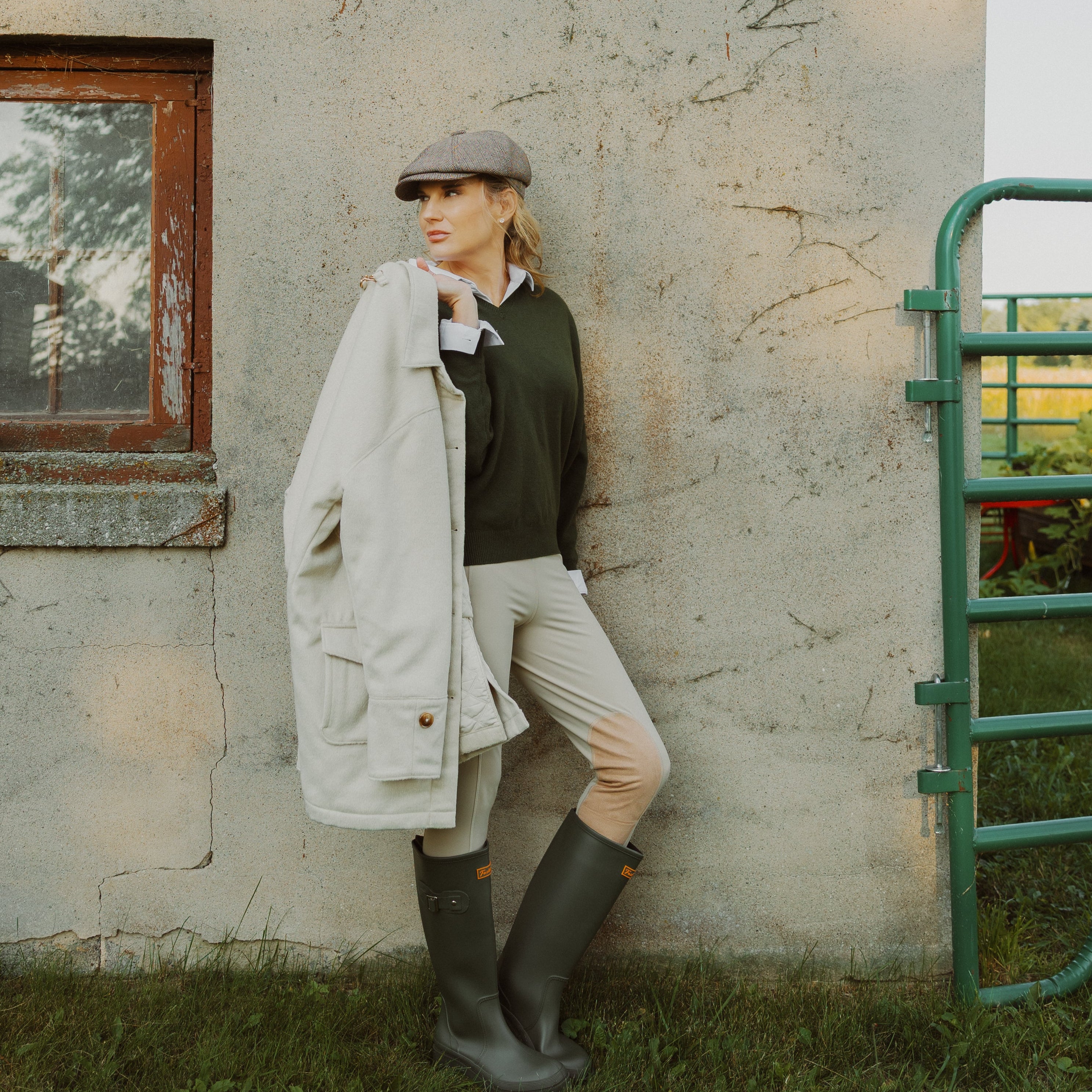 A woman stands outdoors against a weathered barn wall, wearing a light cream coat, dark green top, beige breeches, and dark rubber boots.