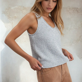 Woman wearing a light blue knitted tank top and beige shorts against a textured wall.