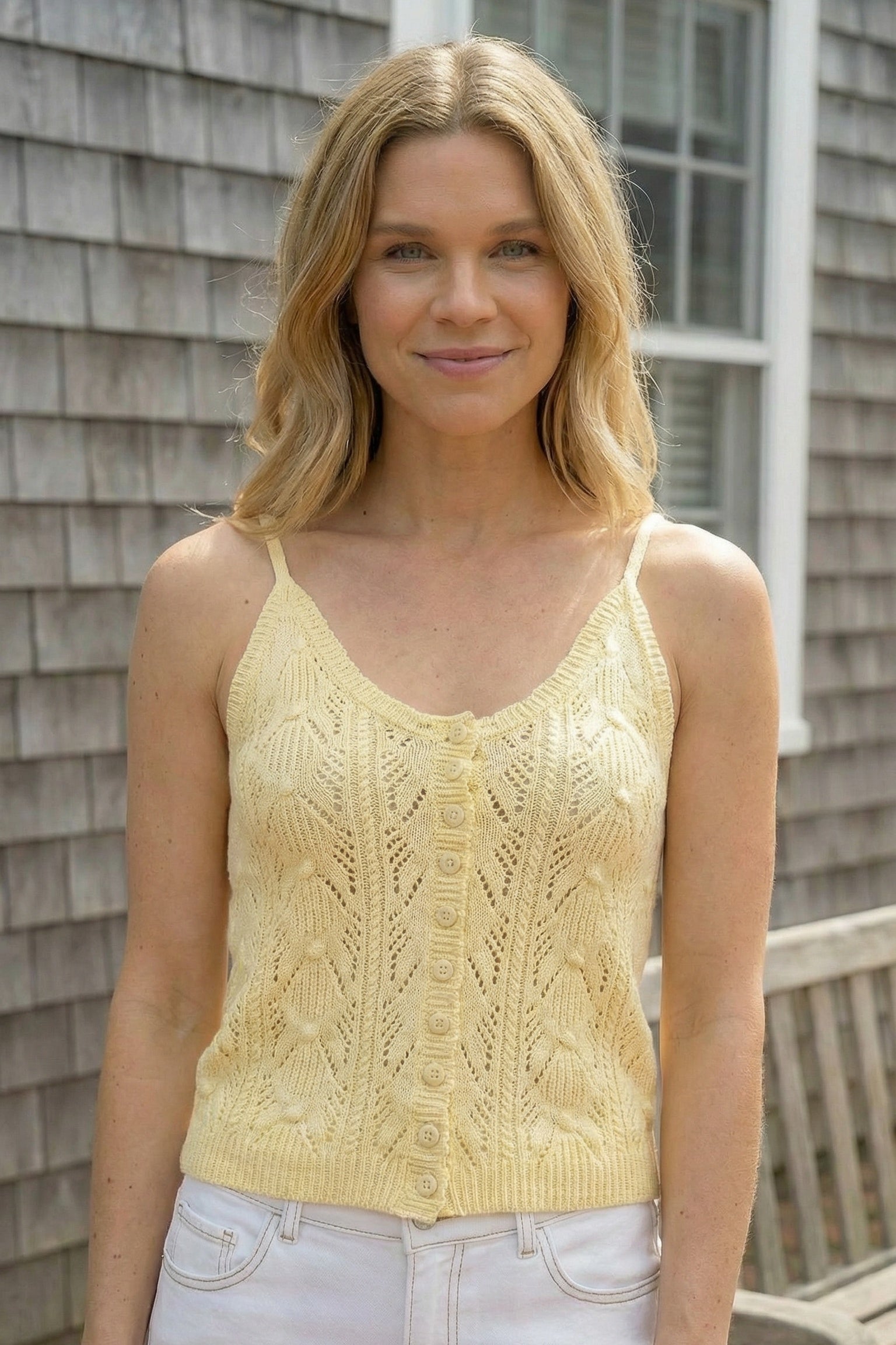 Woman wearing a yellow knitted tank top standing in front of a wooden house.