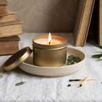 Candle in a gold container on a white surface with books and greenery