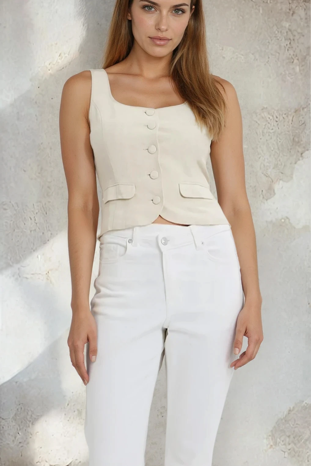 Woman wearing a beige sleeveless top and white pants against a textured wall.