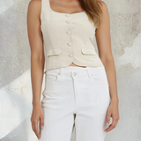 Woman wearing a beige sleeveless top and white pants against a textured wall.