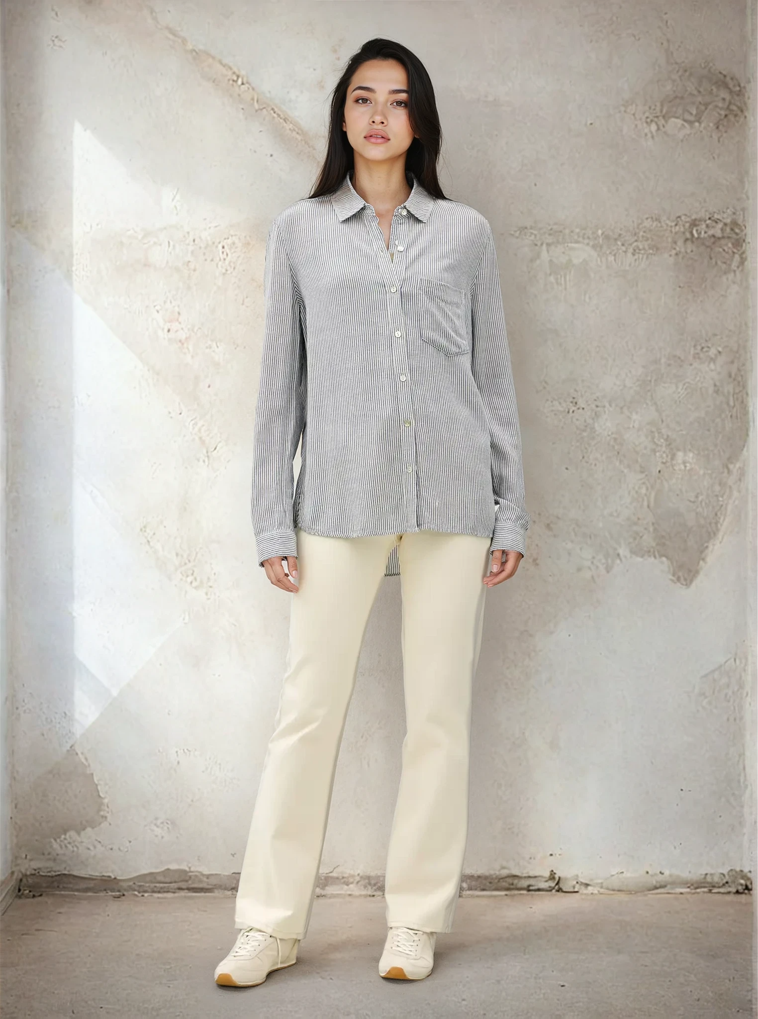 Woman wearing a gray button-up shirt and white pants against a textured wall.