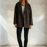 Woman wearing a brown coat, white top, black pants, and brown boots against a neutral background