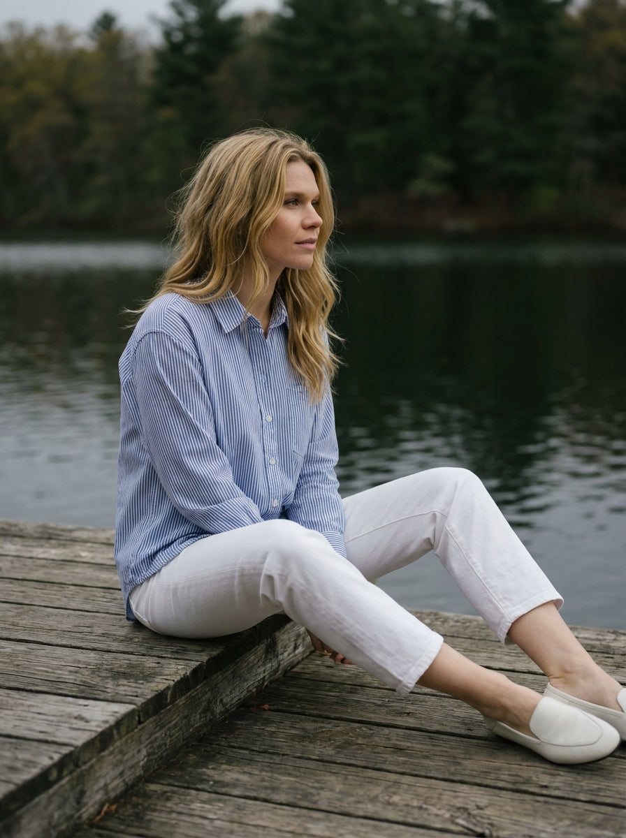 Woman sitting on a wooden dock by a lake wearing a blue striped shirt and white pants.