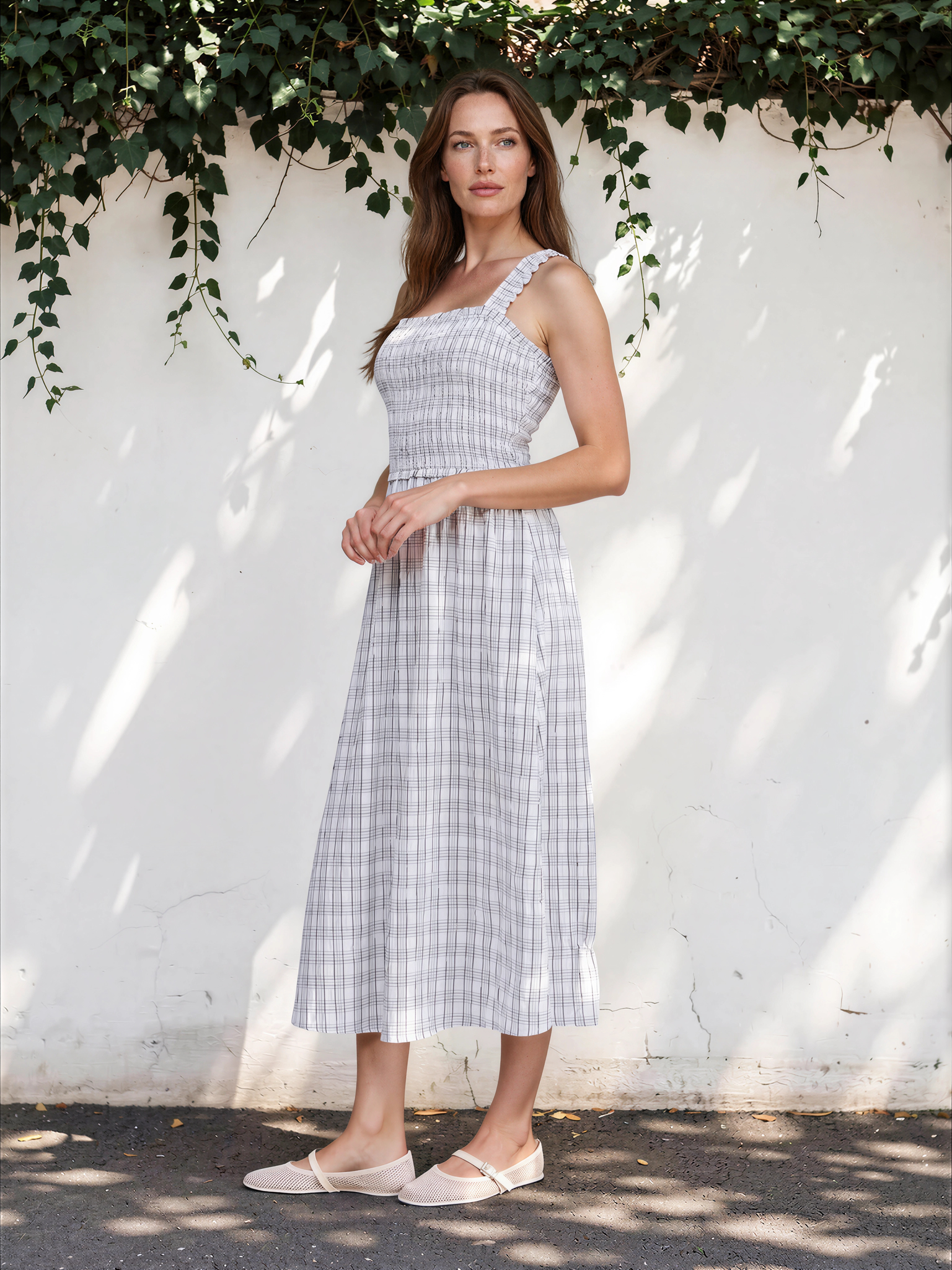 Woman in a checkered dress standing against a white wall with greenery.