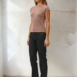 Woman wearing a brown top and black jeans against a white wall.