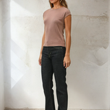 Woman wearing a brown top and black jeans against a white wall.