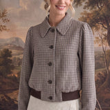 A woman modeling a cropped, long-sleeve button-front jacket with a brown and beige houndstooth pattern, paired with a cream skirt, set against a rustic landscape backdrop.